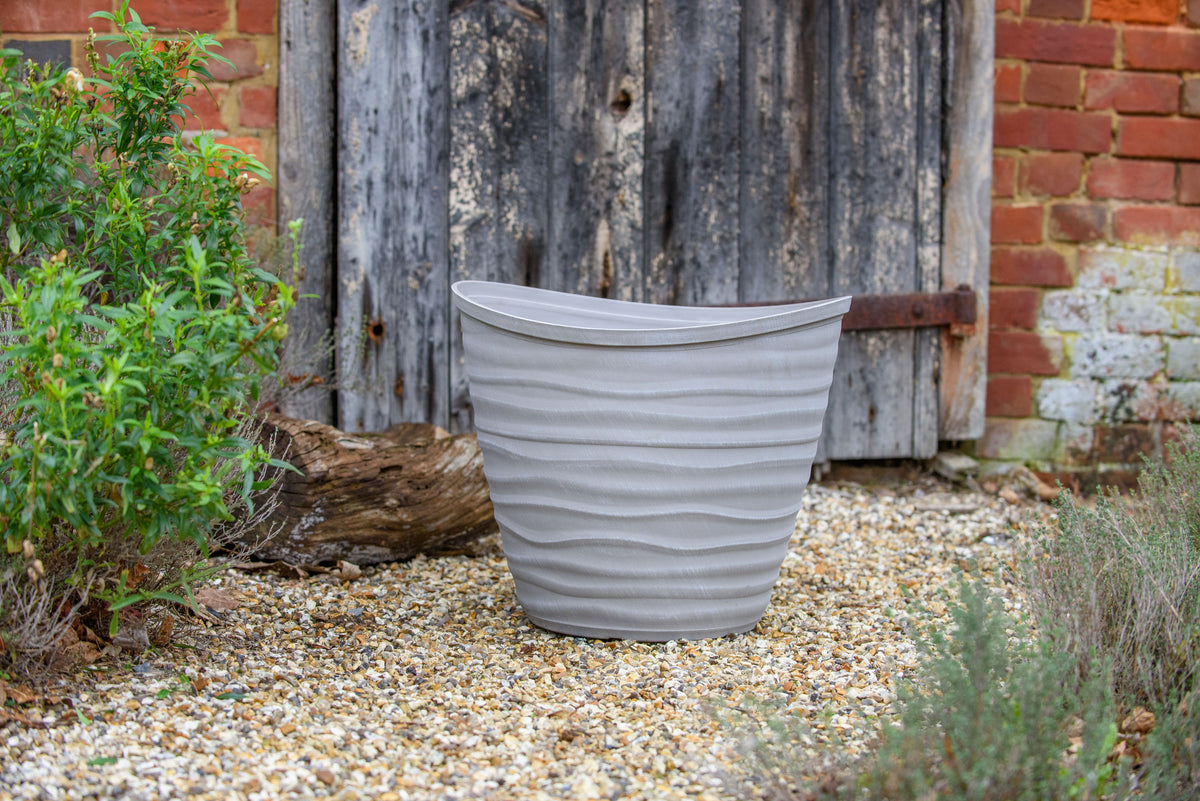 Gray ribbed planter on a gravel surface with a rustic wooden wall and plants in the background