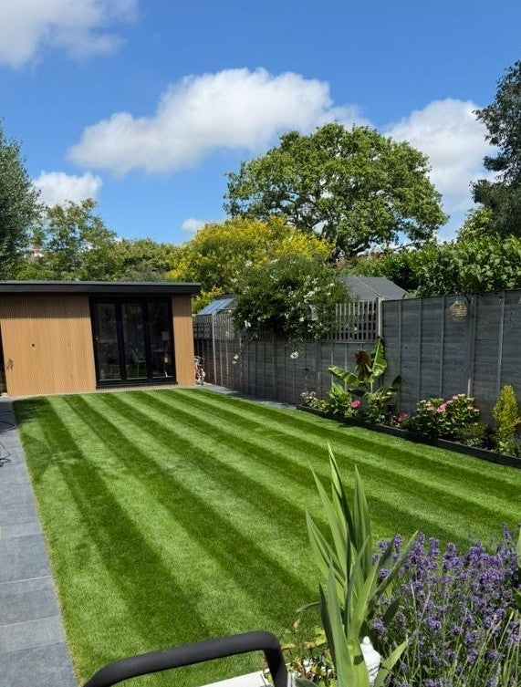 Lush green lawn turf with a garden and wooden shed under a blue sky.