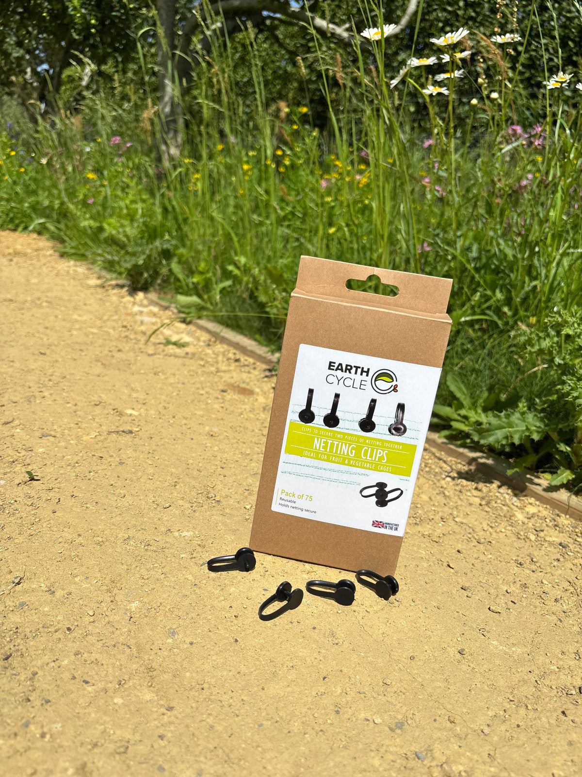 Earth Cycle netting clips in their brown cardboard packaging, placed on a dirt path with a lush green garden background.