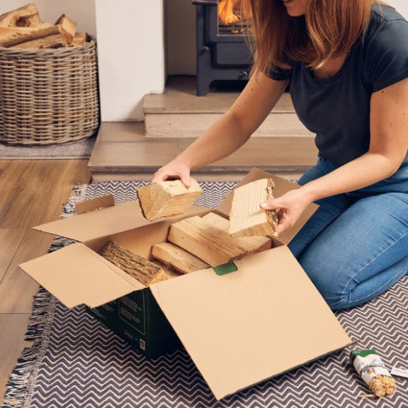 Person unpacking firewood from a cardboard box in a home setting with a fireplace.